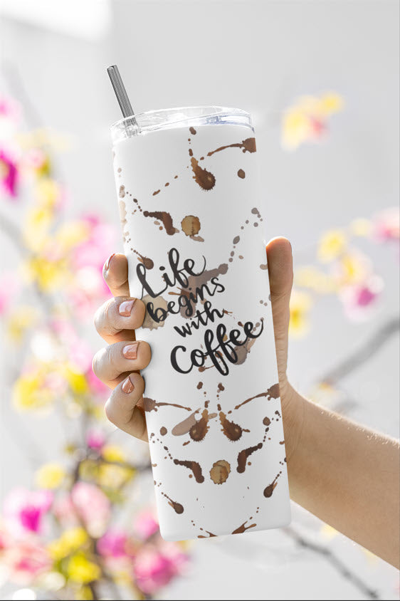 A hand holding a white tumbler with the phrase 'Life begins with coffee' written in a playful script. The tumbler is decorated with brown coffee splatters, giving it a fun and casual look. The tumbler has a clear lid and a metal straw, and is being held in front of a blurred background featuring pink and yellow flowers.